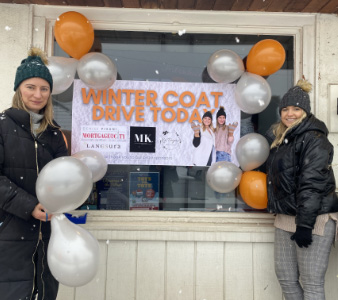2 women in winter coats and toques, standing in front of a window with balloons and a sign that says
										“Winter Coat Drive Today”